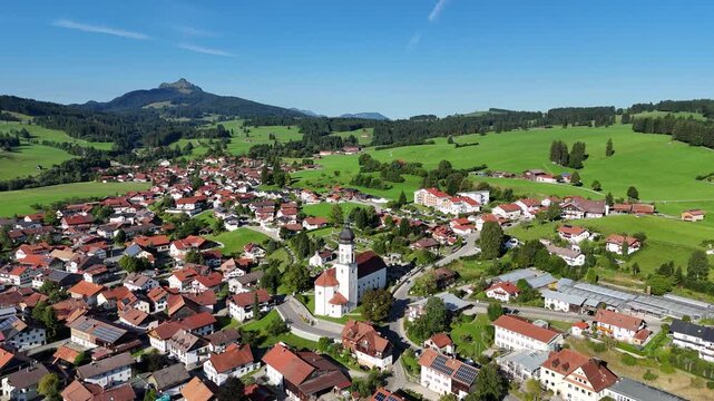 Panorama flight towards parish church St. Ulrich in village Wertach with houses and meadows and mountain summit Gr&uuml;nten in Allg&auml;u Alps, Bavaria, Germany