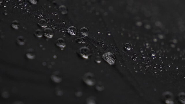 Close up of water droplets splashing and bouncing on a textured black water repellent nylon fabric.