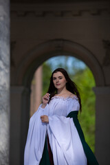 Obraz premium Vertical photo. Young adult woman with bare shoulder, green white medieval dress stands near arch, marble column of palace, looks at camera. Concept of diverse beauty, Middle Ages, pray, magic, faith