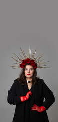 Naklejka premium Vertical photo banner. Young adult white woman with DIY golden halo crown, rose on head, red lips, tulle gloves, black jacket looks at camera on gray. Diverse beauty, pray, faith, fantasy. Copy space