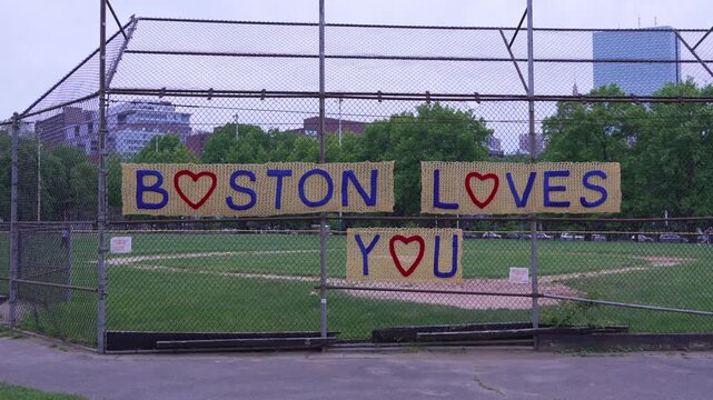 Colorful knitted message reading Boston Loves You attached to chain link fence of empty baseball field in urban park in Boston, USA