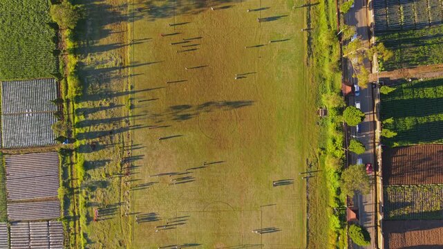 Aerial view of people playing on a grassy soccer field with long shadows near agricultural plots in Dieng Kulon, Batur, Central Java, Indonesia.