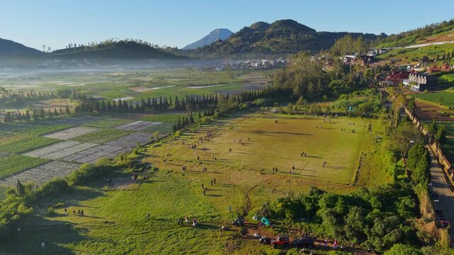 Aerial view of the Dieng Kulon field with people walking near agricultural plots and distant mountains under a clear sky in Batur, Central Java, Indonesia.