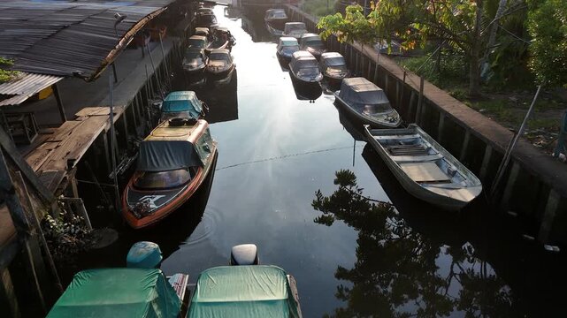 A drone view showing a narrow urban canal lined with motorboats moored on both sides, reflecting the morning sunlight on the dark, calm water, in west borneo, Indonesia.
