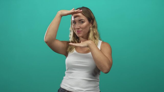 Woman blonde in white tank top framing her face with palms, hands forming a rectangle above forehead and below chin in studio; playful curiosity.