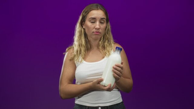 Woman holding milk bottle in bare hands, tilting and examining blue cap with a doubtful pout against a purple studio backdrop; skeptical hesitation.