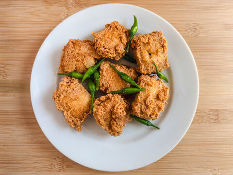 Crispy fried tofu or tahu crispy in cube pieces on a white plate at wooden table. green hot chili peppers garnishes the dish, adding color. Simple, appetizing presentation. Indonesian street food
