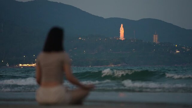 Woman meditating on beach shoreline with sea waves and Lady Buddha in Da Nang, Female practicing mindfulness by ocean at sunset with coastal mountains 4k