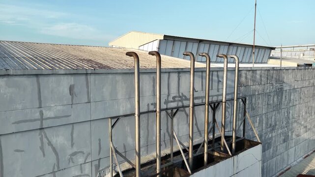 A drone shot showing a row of industrial metal exhaust pipes on the exterior wall of a factory building, with a corrugated roof in the background.