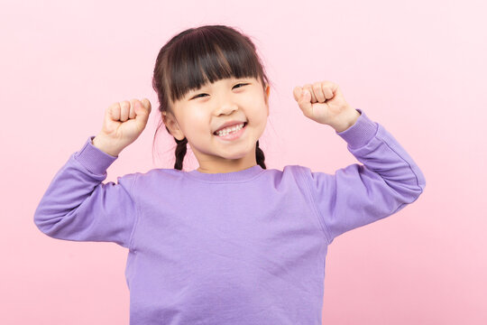 Excited little Asian girl raising fists in victory celebrating success on a pink background.