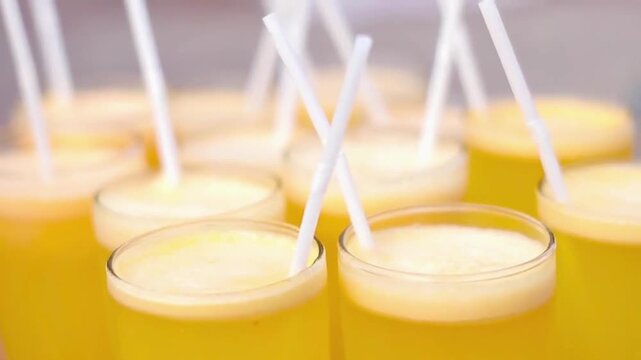 Rows of freshly squeezed orange or pineapple juice glasses with white straws are prepared for a catering event buffet