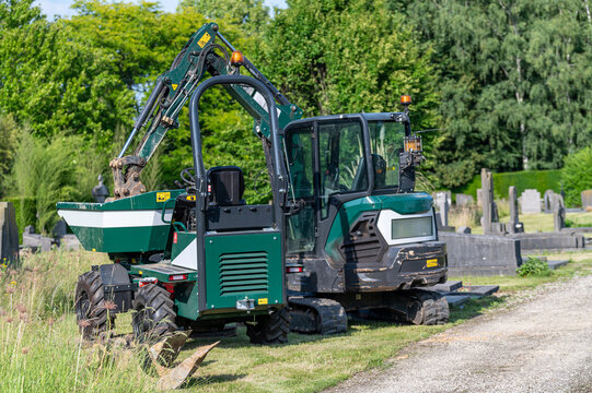 Small green compact excavator and tracked dumper parked at a cemetery for maintenance work.Mini digger and utility vehicle construction machinery operating on a graveyard background.