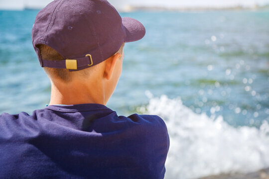 Caucasian Teen Boy in Navy Cap Back View Looking at Blue Sea Splashing Wave Summer Vacation Closeup