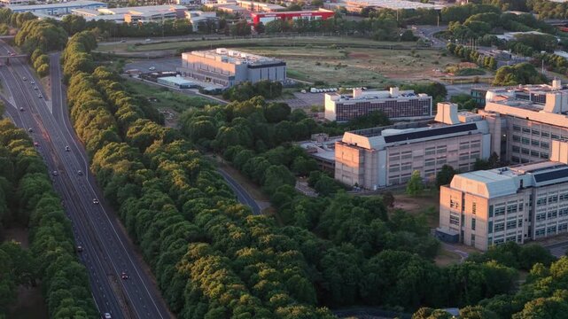 Aerial view of the GSK Stevenage pharmaceutical campus and the A1 motorway during golden hour with lush green trees and traffic in Stevenage, England, United Kingdom.