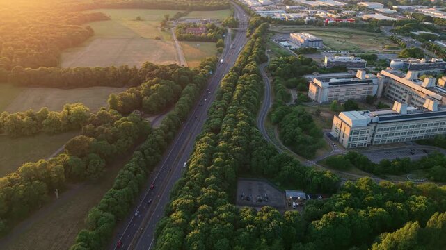 Aerial view of the GSK Stevenage research campus and a major motorway surrounded by lush green woodland during golden hour in Stevenage, England, United Kingdom.