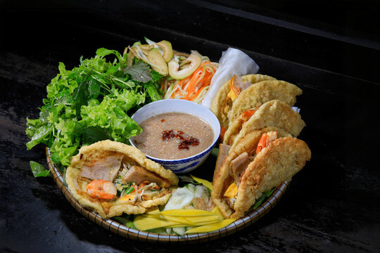 A traditional Vietnamese dish called Banh Khoai, a central region specialty. Deep-fried crispy pancakes filled with shrimp, pork, and bean sprouts, served on a round bamboo platter alongside a rich pe