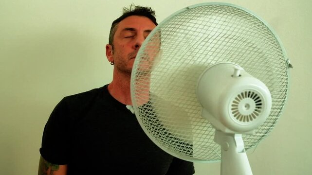 A man enjoys the coolness of the fan up close to counteract the summer heat. The man in the video is forty-two years old, is an Italian of Caucasian origin and wears a dark gray T-shirt