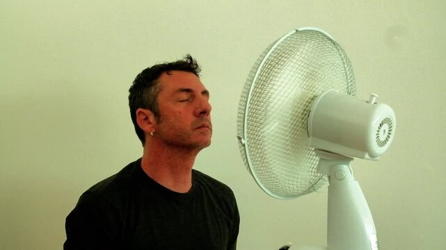 A man enjoys the coolness of the fan up close to counteract the summer heat. The man in the video is forty-two years old, is an Italian of Caucasian origin and wears a dark gray T-shirt