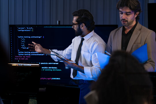 South Asian developer gestures toward a Python code display on a large screen in a dark briefing room while a Caucasian colleague holds documents beside him.