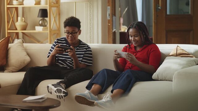 Happy Black siblings playing online video games on their phones, enjoying fun