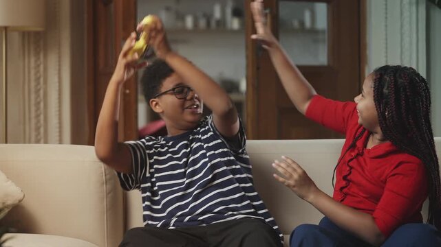 Happy Black siblings laughing and fighting over a video game controller at home