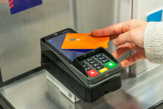 Close-up of a customer making a contactless payment with a credit card at a checkout terminal.Person paying with an orange debit card on a POS terminal device. Retail shopping and wireless technology 