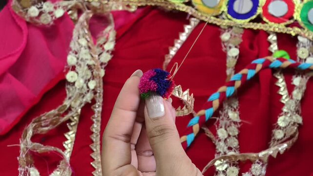 Close-up of hands stitching a colorful pompom embellishment onto a handcrafted costume, showcasing traditional textile craftsmanship, embroidery techniques, and artisan fashion detailing.