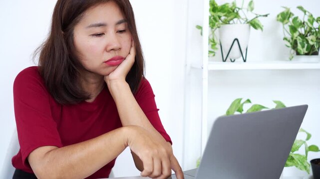 Asian woman looking bored, thoughtful, or unmotivated while working on a laptop at a bright home office desk, representing procrastination