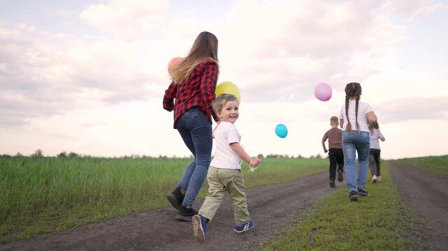 Small group run down long dirt road close to green field. Active child hold colorful balloon. Happy kid play outdoor in beautiful nature. Group run very fast. Child play with bright balloon.
