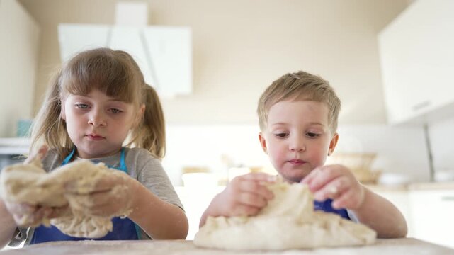 Children kneading dough in modern white kitchen at home. Boy and girl in blue aprons play with flour dough on counter. Siblings enjoy baking together. Kids learn cooking during fun family activity.