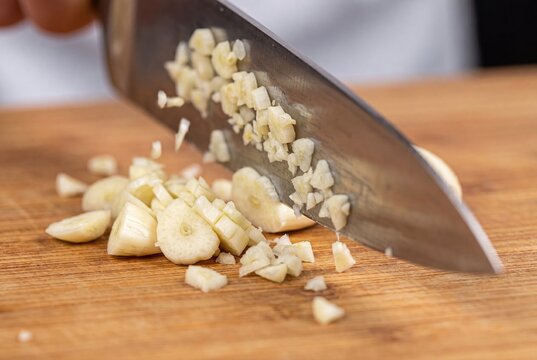 Close-up of a chef's knife finely mincing fresh garlic cloves on a wooden cutting board for cooking.