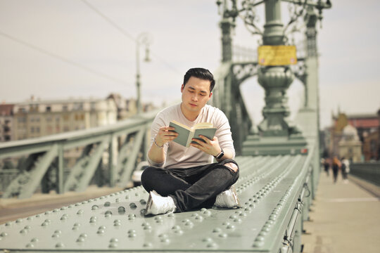 Young Asian man reading a book on a historic bridge
