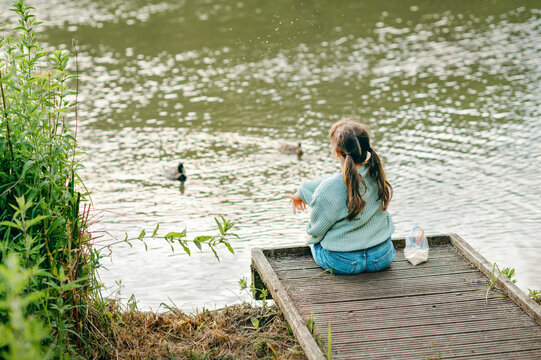 Child girl sitting on lakeside dock feeding birds on a calm pond