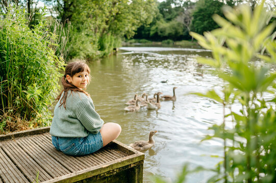 Child girl looking back while sitting beside a lake with waterfowl