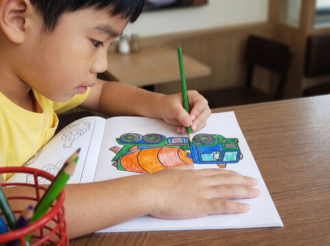 Close up side view of cute Asian boy focusing on coloring a cement mixer truck book with a green pencil while sitting at a wooden table in a cafe