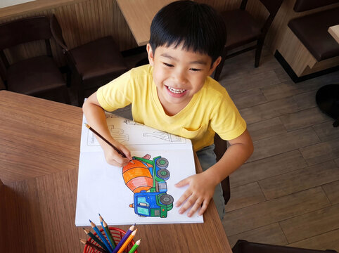 High angle view of happy cute Asian boy laughing cheerfully while holding a pencil to color a cement mixer truck drawing book on wooden table at restaurant