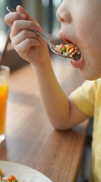 Extreme close up of cute Asian boy hand holding a spoon to eat healthy salmon fried rice with vegetables at mouth level on wooden table