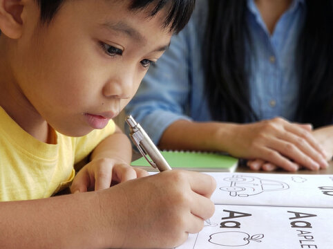 Close up side view of concentrated cute Asian boy practicing writing English alphabet Aa in exercise book with his mother supporting him closely at home