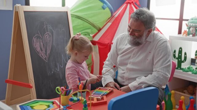 Man watches and guides little girl playing xylophone at a colorful playroom table with chalkboard heart and toys; intergenerational bonding warmth.