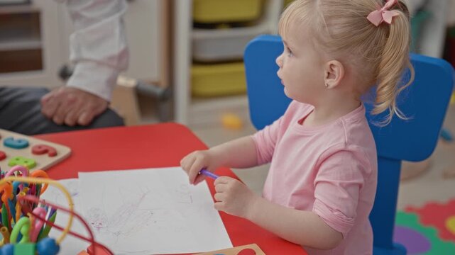 Man drawing with purple crayon on paper in classroom while toddler girl sits with arms crossed at a table; learning frustration.