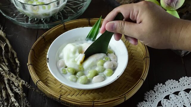 Scooping traditional Thai dessert Bua Loy Kaew, translucent tapioca balls with taro, orange sweet potato, and pumpkin in warm coconut milk soup, served in a ceramic bowl on a bamboo tray.