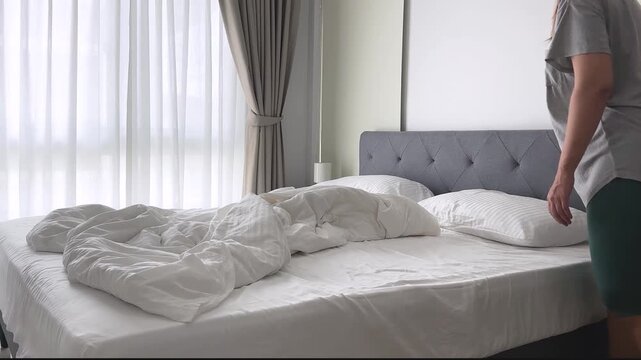 Woman making a bed with white bedding in a bright bedroom, everyday housekeeping and home routine in a modern apartment interior.