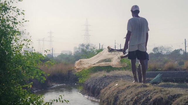 Daylight outdoor shot of two Indian Asian male fishermen casting a fishing net in the pond, lake, or canal water in search of fish on a sunny but windy day. 