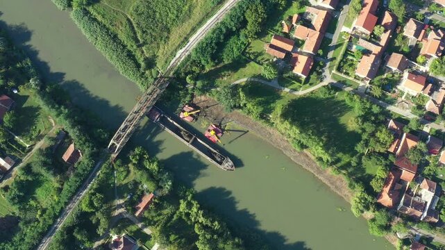 Above top view on two excavators dredge as they dredging, working on river, canal, deepening and removing sediment, mud from riverbed in a polluted waterway near old railroad bridge.