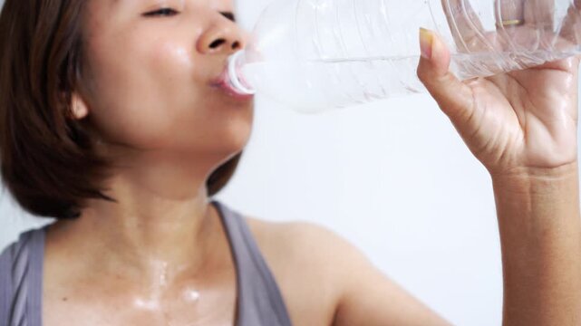 sweaty Asian woman drinking fresh water from a bottle after a workout to relieve thirst, wiping her forehead while feeling tired and exhausted from hot weather at home