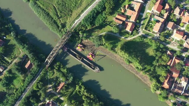 Above top view, dolly move downward over two excavators dredge as dredging, work on river, canal, deepening and removing sediment, mud from riverbed in a polluted waterway near old railroad bridge.