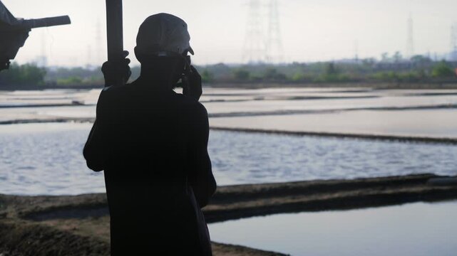 A daylight outdoor silhouetted shot of a male worker or labourer wearing a cap standing in a hut, talking on a mobile phone or smartphone against the salt pan flats filled with water on a sunny day 