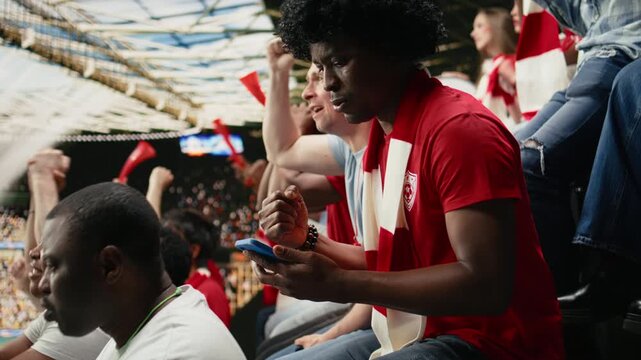 Black football fan with wig monitor live betting odds at stadium during live soccer match filled with cheering and crowd excitement.