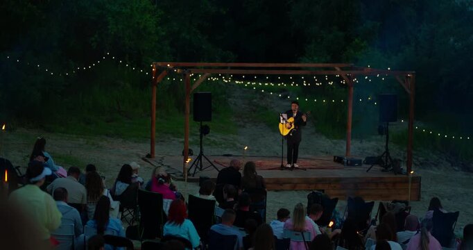 Solo acoustic guitarist performing on a string-lit pergola stage at dusk