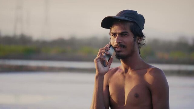 Daylight outdoor shot of a shirtless happy Indian Asian young teenage worker wearing a cap talking on a mobile phone or smartphone with salt pan flats visible in the background in the evening. 